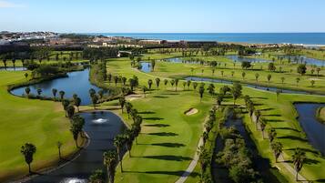 Ein Golfplatz mit mehreren Wasserhindernissen, umgeben von Grünflächen und Palmen, mit Blick auf den Ozean im Hintergrund.