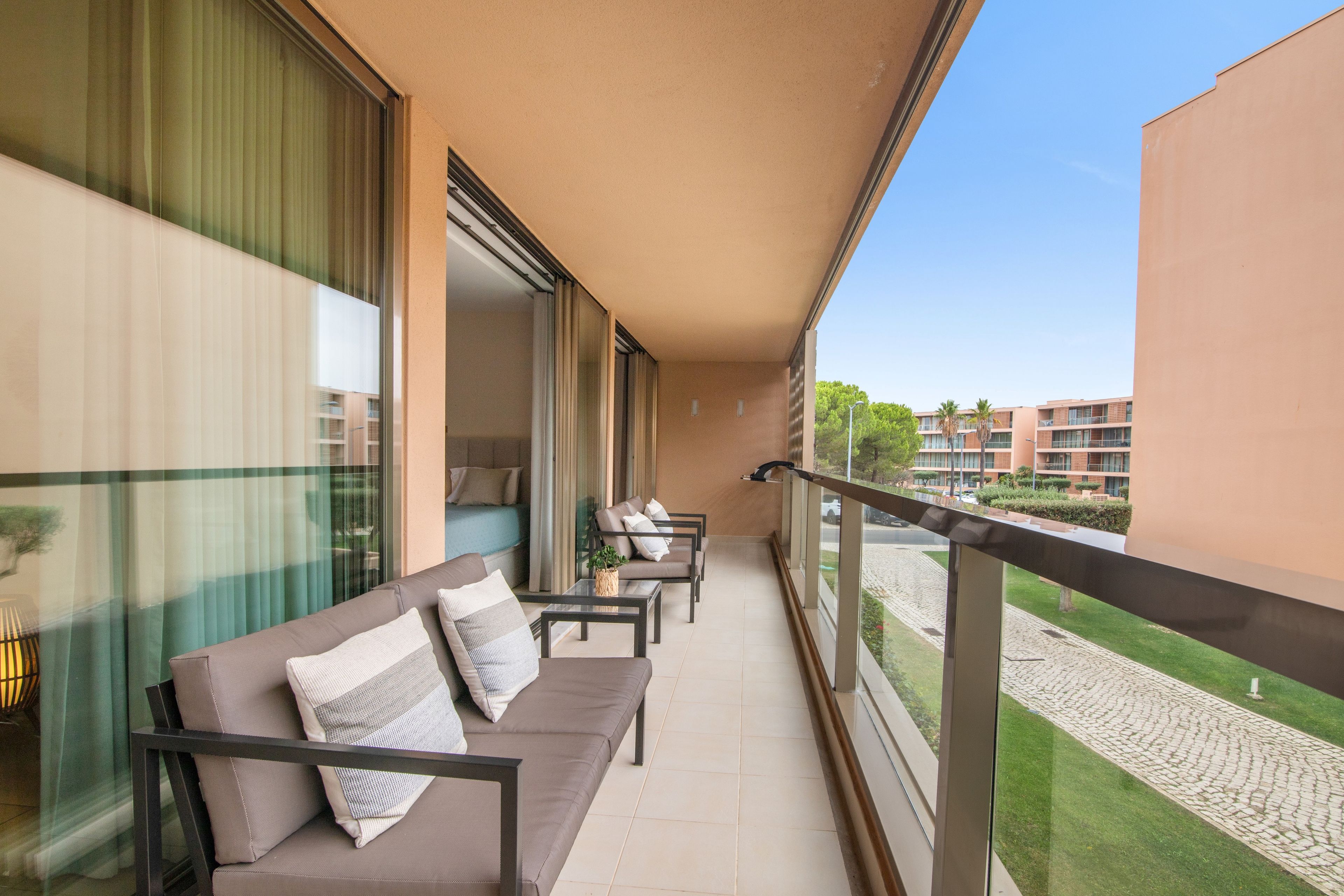 Un balcón con sofá y sillones, una mesa de vidrio y vistas a un conjunto de edificios.