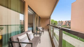 Un balcón con sofá y sillones, una mesa de vidrio y vistas a un conjunto de edificios.