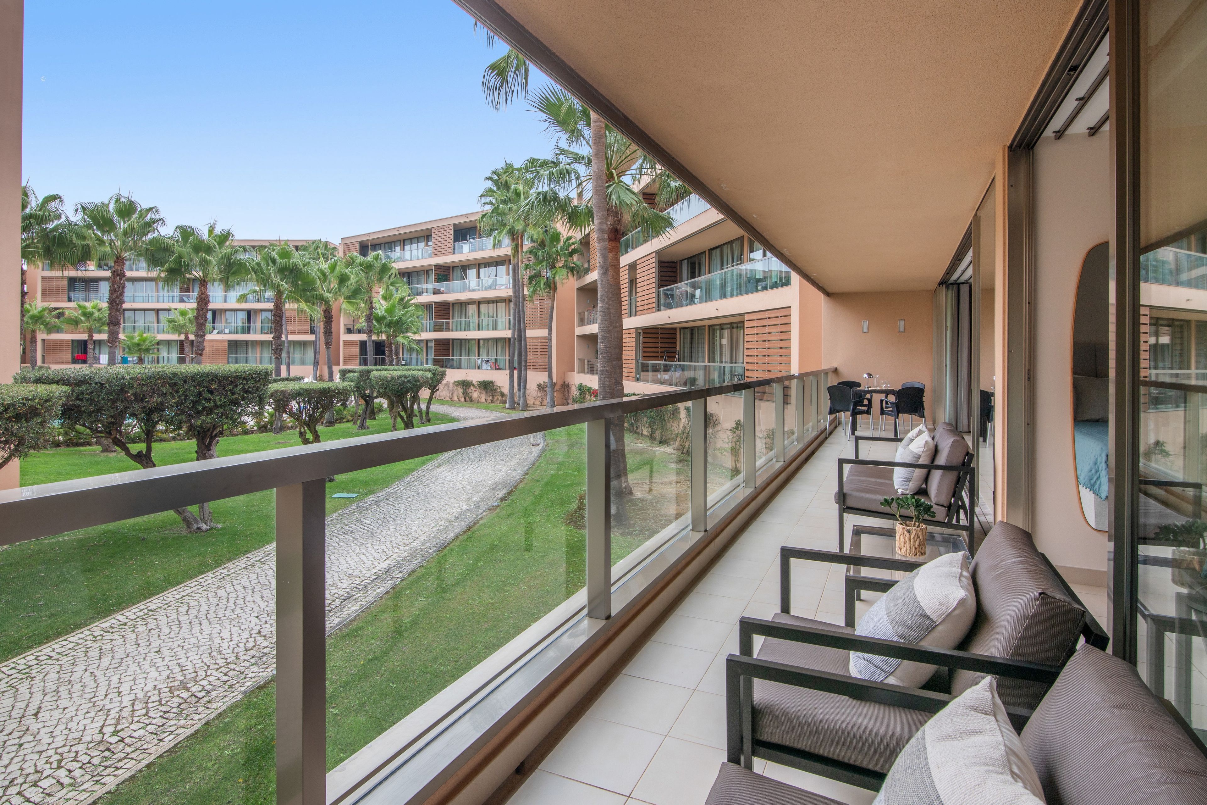 A balcony with a seating area, a view of a courtyard with palm trees, and a building with multiple balconies.