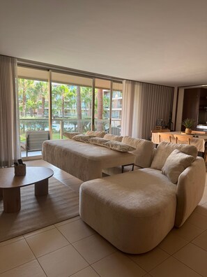 Habitación de hotel moderna con una cama grande, un sofá, una mesita y vistas a una piscina y palmeras.