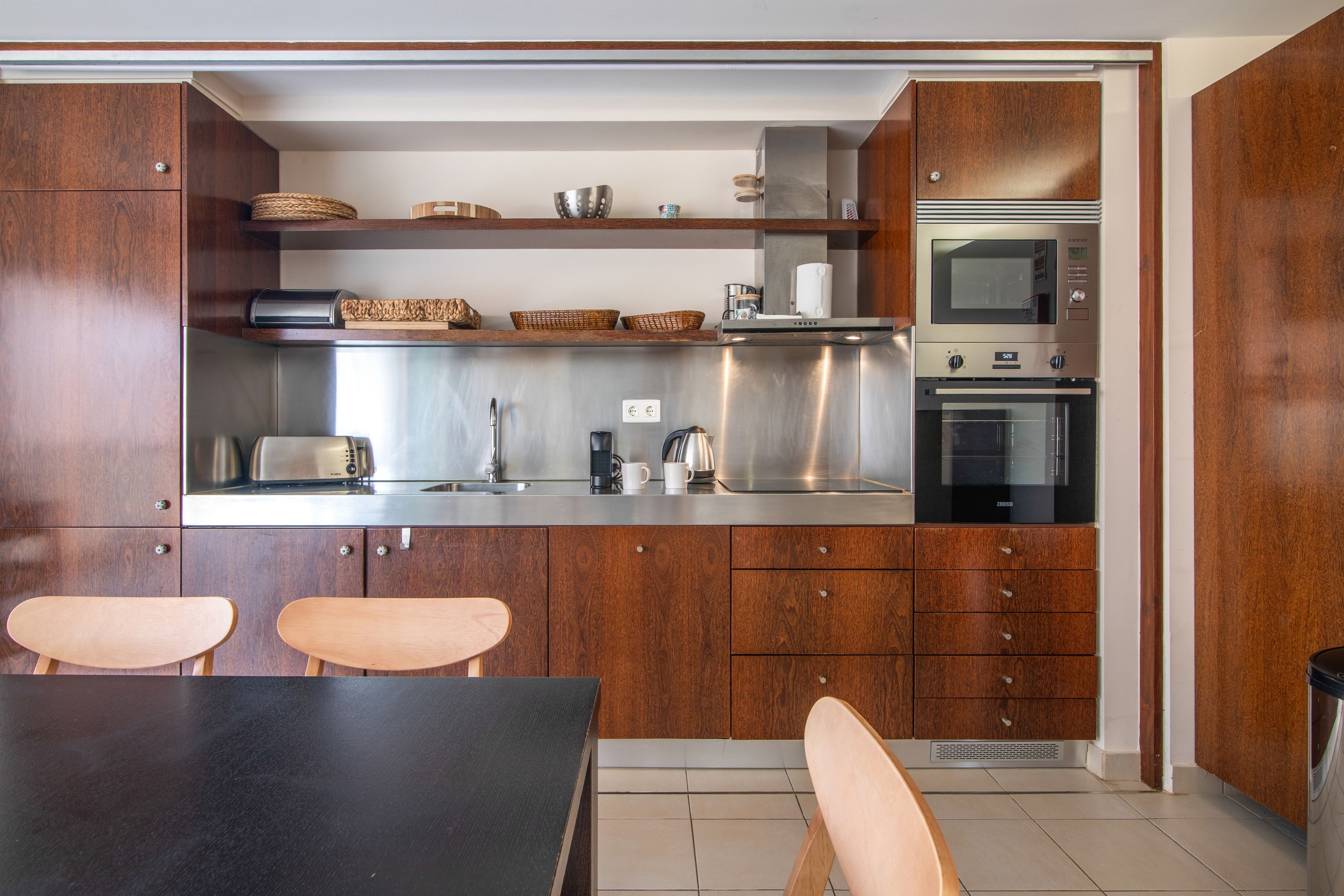 Een moderne keuken met houten kastjes, roestvrijstalen apparatuur en een eethoek met twee stoelen.
