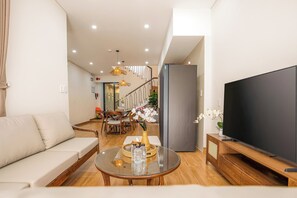 A modern living room with a sofa, a coffee table with bottles, a TV, and a staircase leading to another level. - The Querencia Arena Private Pool villa and Free Pick up (Đà Nẵng)