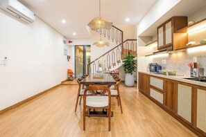 A modern kitchen with wooden cabinets, a dining area with a wooden table and chairs, and a staircase with a unique railing design. - The Querencia Arena Private Pool villa and Free Pick up (Đà Nẵng)