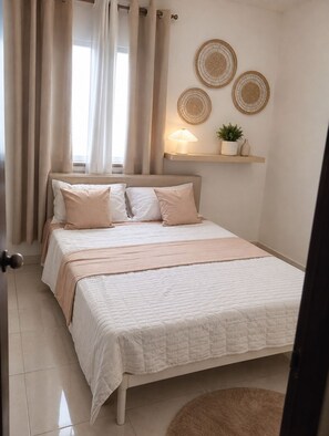 A bedroom with a bed, curtains, a wall-mounted shelf with decorative items, and a door. - Serenity Cove, A  3-bedroom apartment with AC in Terrezas del Este (Villa Hermosa)