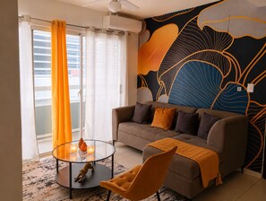 Een woonkamer met een bank, fauteuil, glazen salontafel en een wand met bladmotief.