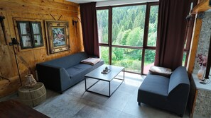 A room with a wooden interior, a sofa, a coffee table, and a view of a forest through large windows. - Vrbo Property (Gornji Orašac)