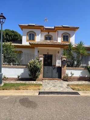 Una casa de dos pisos con techo de tejas, entrada protegida por una reja y un pequeño jardín con palmeras.