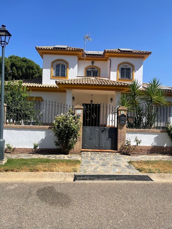 A two-story house with a tiled roof, a gated entrance, and a small garden with palm trees.