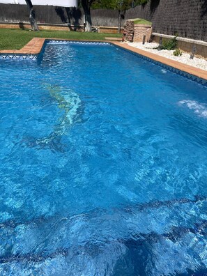 Piscina rectangular con agua cristalina azul, rodeada por un muro de piedra y una zona verde.