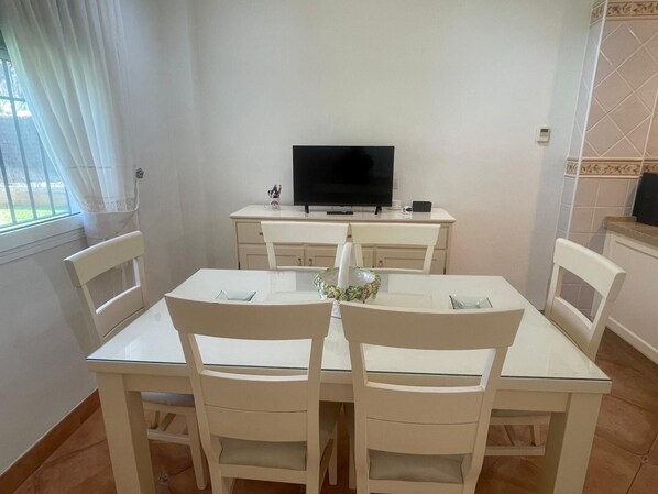 Un comedor con una mesa y sillas, un televisor sobre un mueble bajo, y una ventana con cortinas.