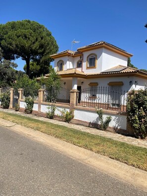 A two-story house with a tiled roof, a fenced yard, and a large tree in the background.