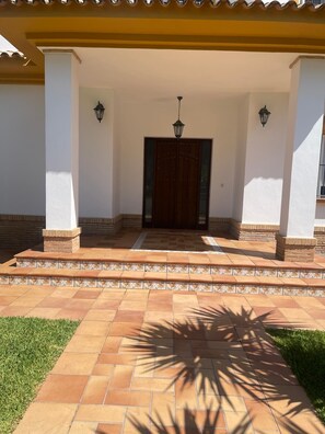 Un acceso cubierto con un sendero adoquinado que lleva a una puerta de madera, flanqueado por dos columnas y luces fijadas a la pared.