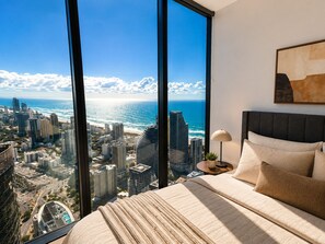 A bedroom with a large window offering a city view, a bed with a headboard, and a bedside table with a lamp. - Horizon Haven - Panoramic Ocean Views (Broadbeach)