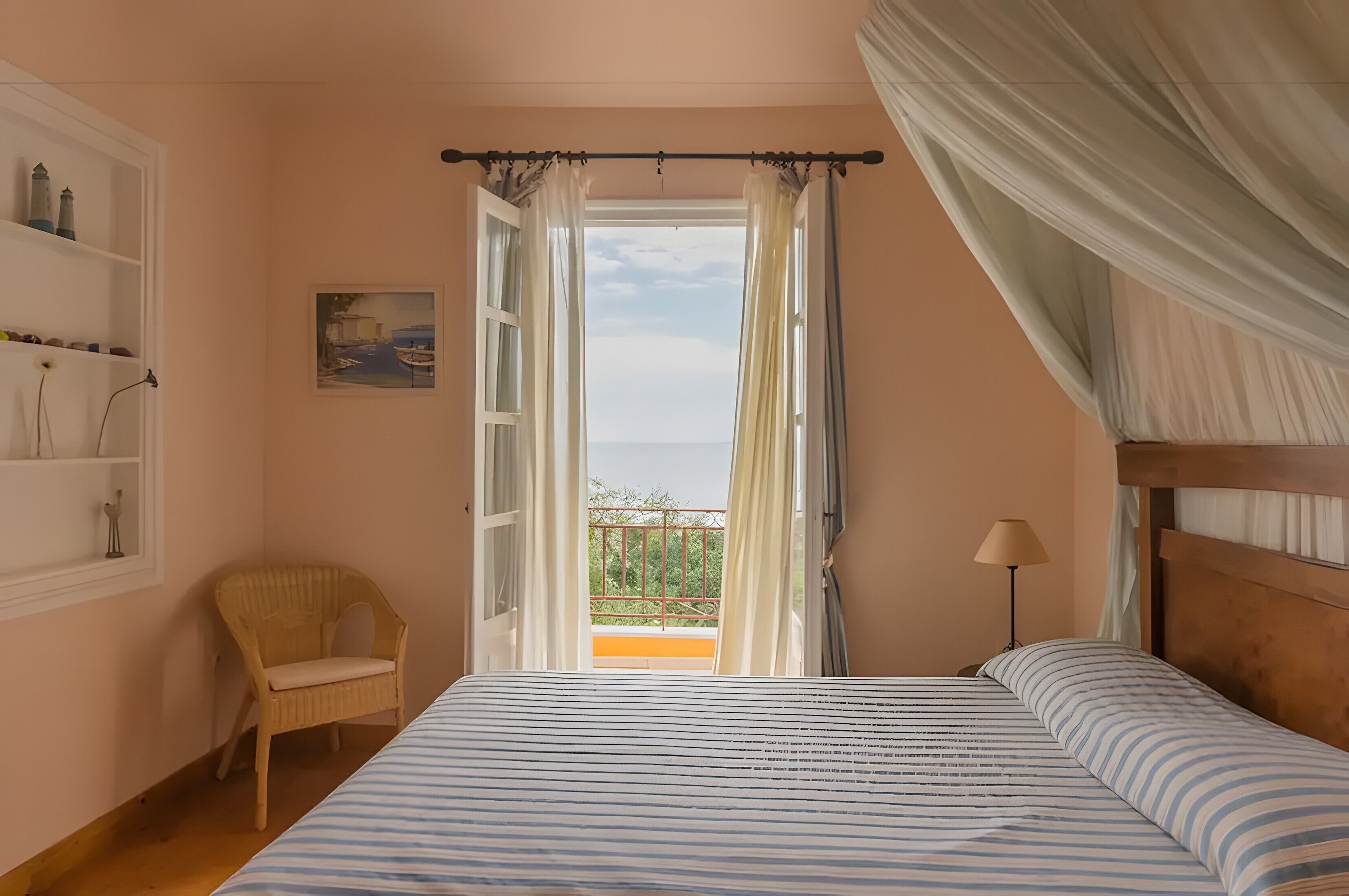 A bedroom with a bed, a chair, a window with curtains, a painting on the wall, and a balcony with a railing.