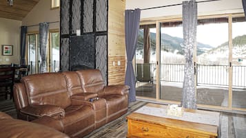 A cozy living room with a leather sofa, a wooden coffee table, and a large sliding glass door leading to a balcony with a mountain view.