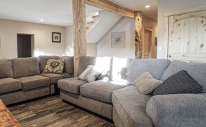 A living room with a sectional sofa, wooden beams, a staircase, and a door with a rustic finish.