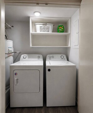 A compact laundry room with two front-loading washing machines, a white shelf above, and a water heater to the left. - Bright & Comfortable Retreat - 2BR Apartment (Waxahachie)
