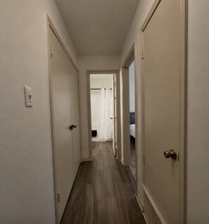 A narrow hallway with wooden flooring and white walls, leading to a room with a bed and a window.