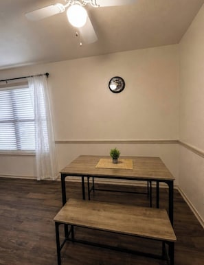 A dining area with a wooden table and bench, a ceiling fan, a clock on the wall, and a window with blinds.