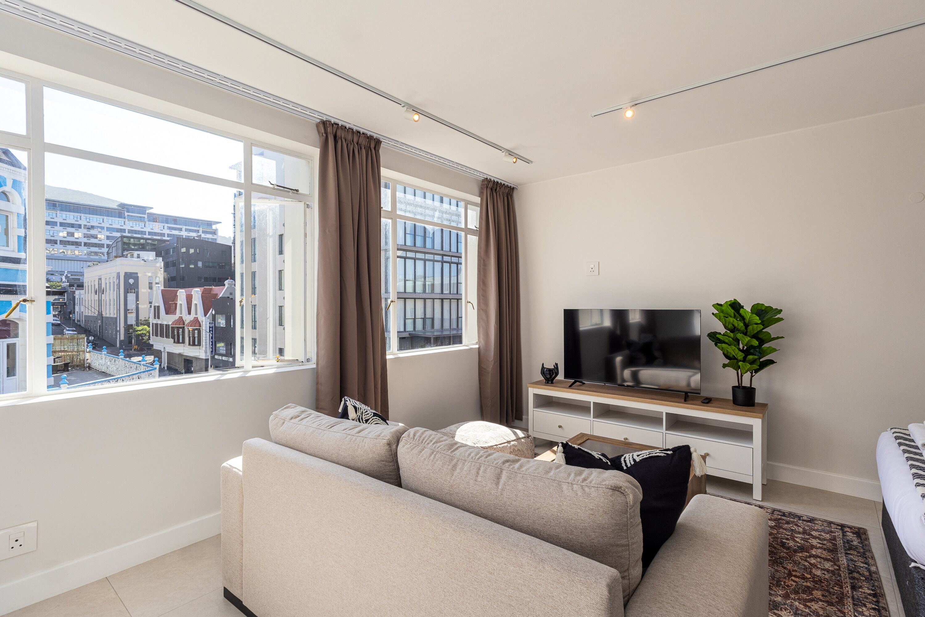 A modern living room with a sofa, TV, and a view of buildings through large windows.