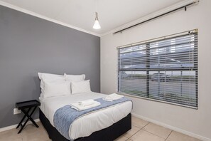 A bedroom with a bed, a bedside table, a hanging light fixture, and a window with blinds. - Spacious 3BR Garden Pool Gym Parking by Ctha (Cape Town)