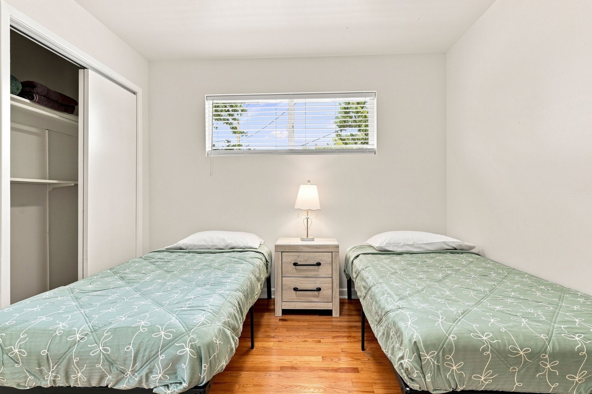 Un dormitorio con dos camas individuales, una mesita de noche con cajones, una ventana con persianas y un armario con ropa colgada.