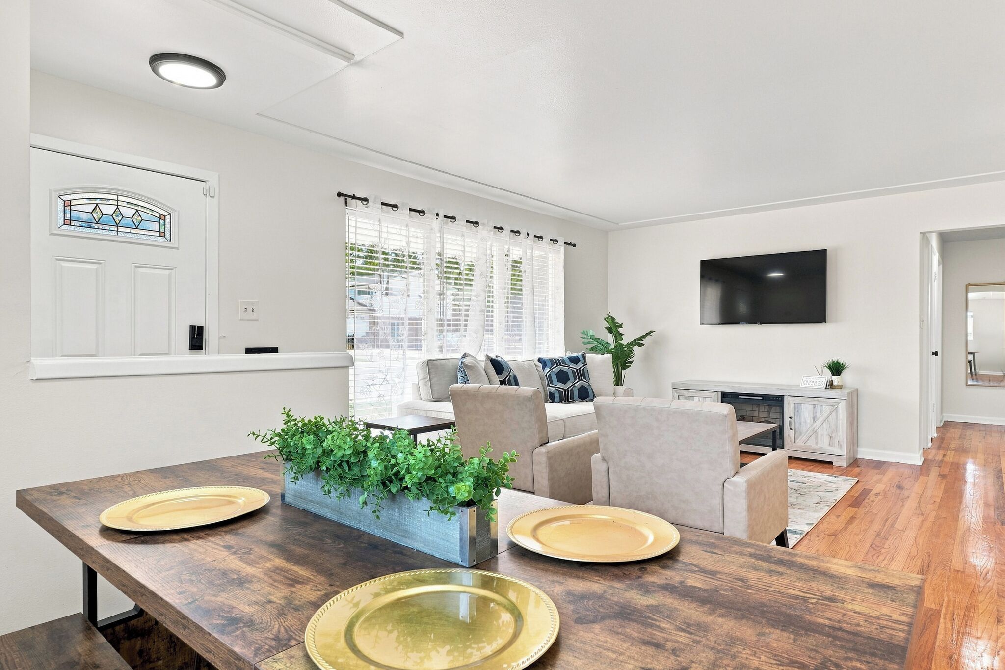 A modern living room with a dining area, featuring a sofa set, a flat-screen TV, and a dining table with plates and a plant centerpiece.
