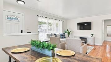 A modern living room with a dining area, featuring a sofa set, a flat-screen TV, and a dining table with plates and a plant centerpiece.