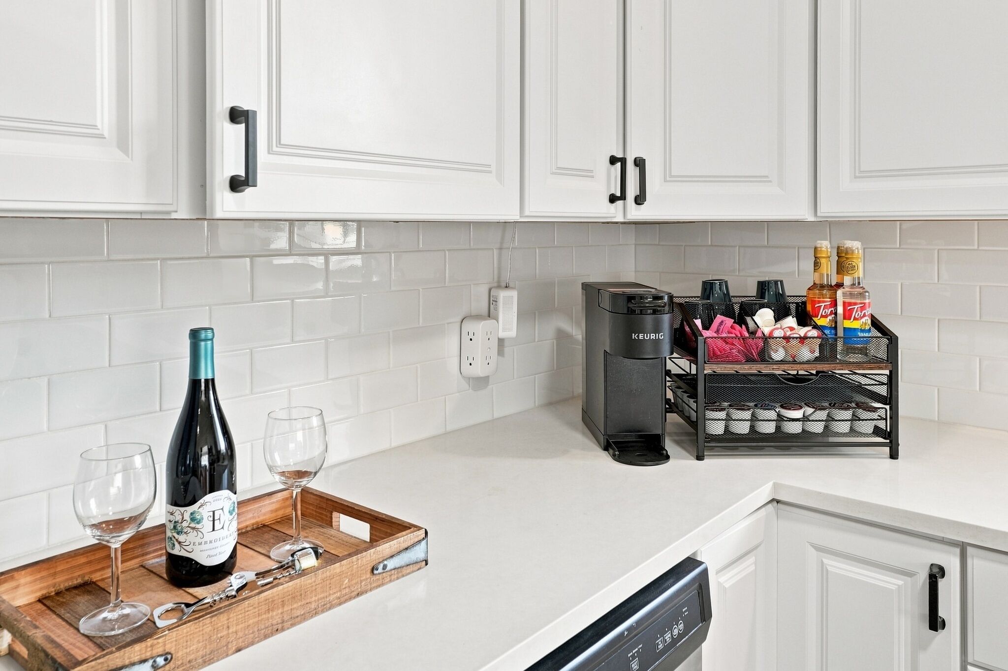 A kitchen with white cabinets, a coffee maker, a wine bottle, and a tray with glasses.