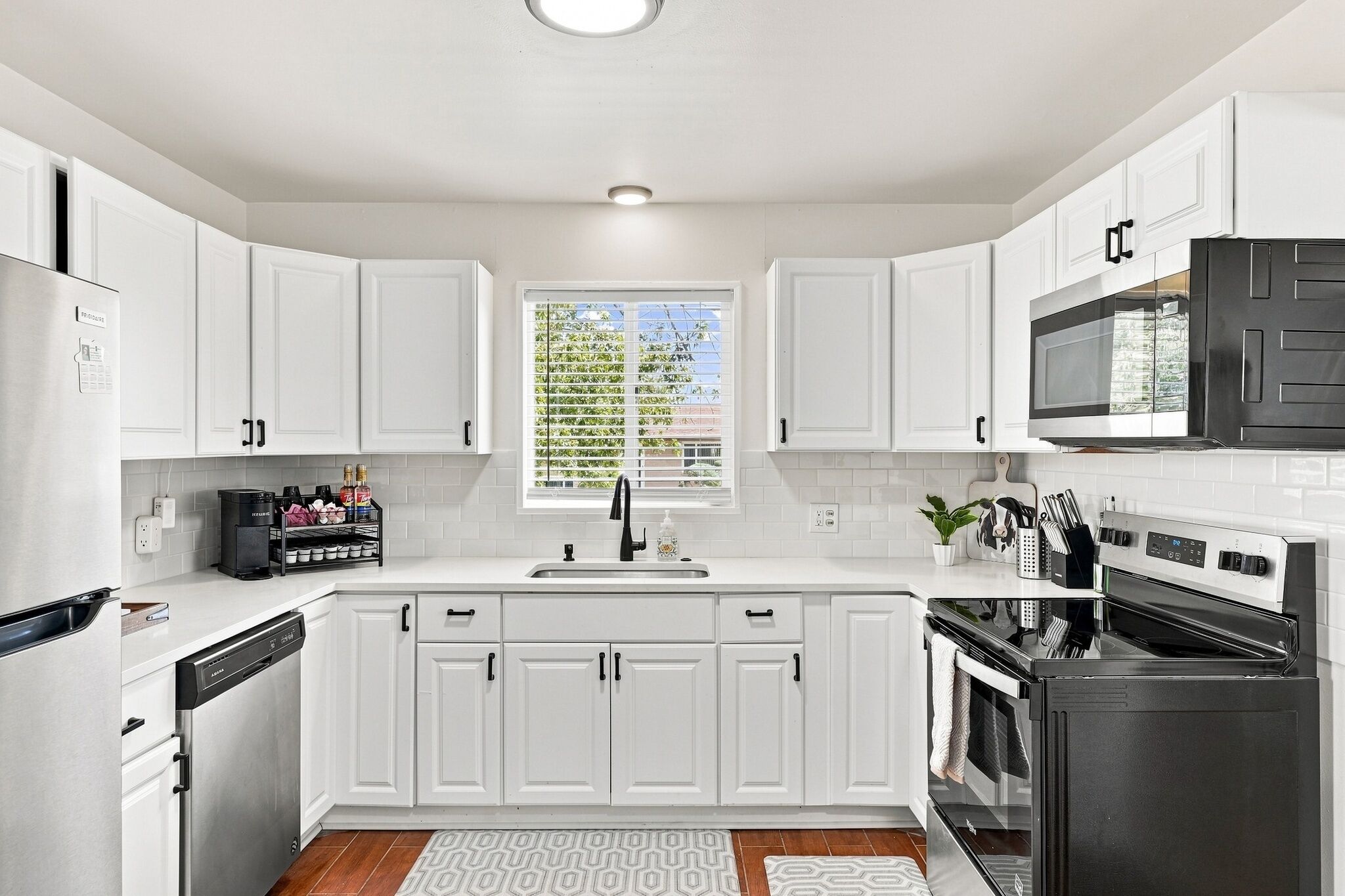 A modern kitchen with white cabinetry, a built-in microwave, a dishwasher, and a large window with blinds.