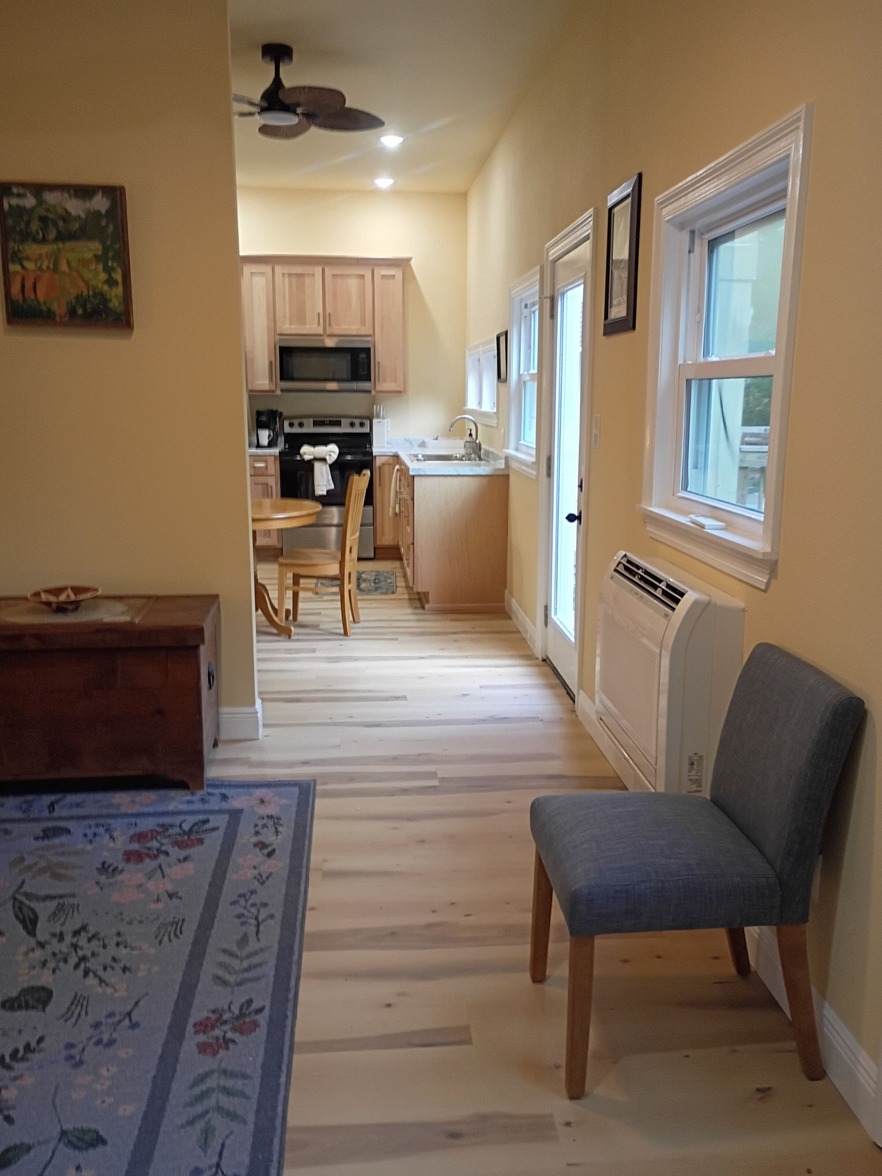 A room with a wooden floor, a blue chair, a wooden table, a kitchen area with cabinets and appliances, and a window with a view.