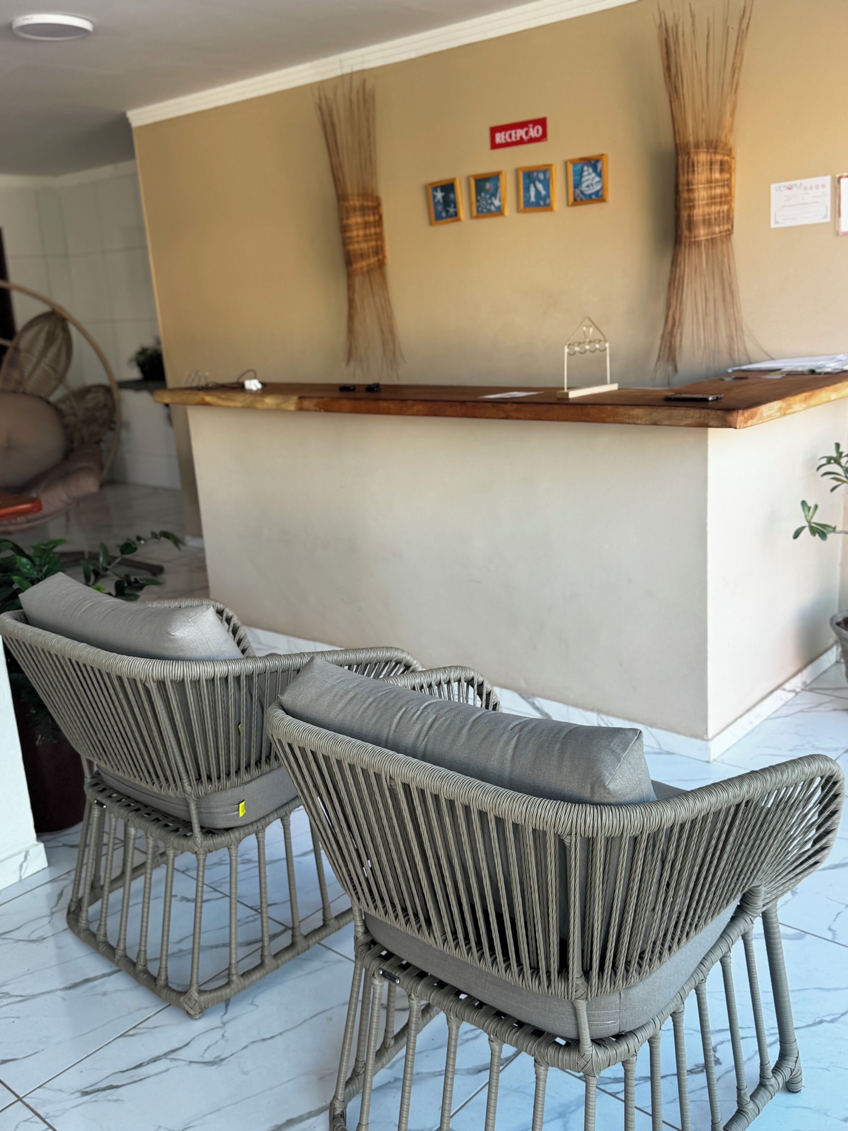 Un espace d’accueil comprenant un comptoir de réception, deux fauteuils en osier avec coussins et un mur orné de tableaux encadrés.