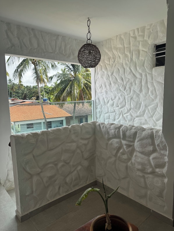 A room with textured white walls, a hanging light fixture, a potted plant, and a view of palm trees and buildings outside.