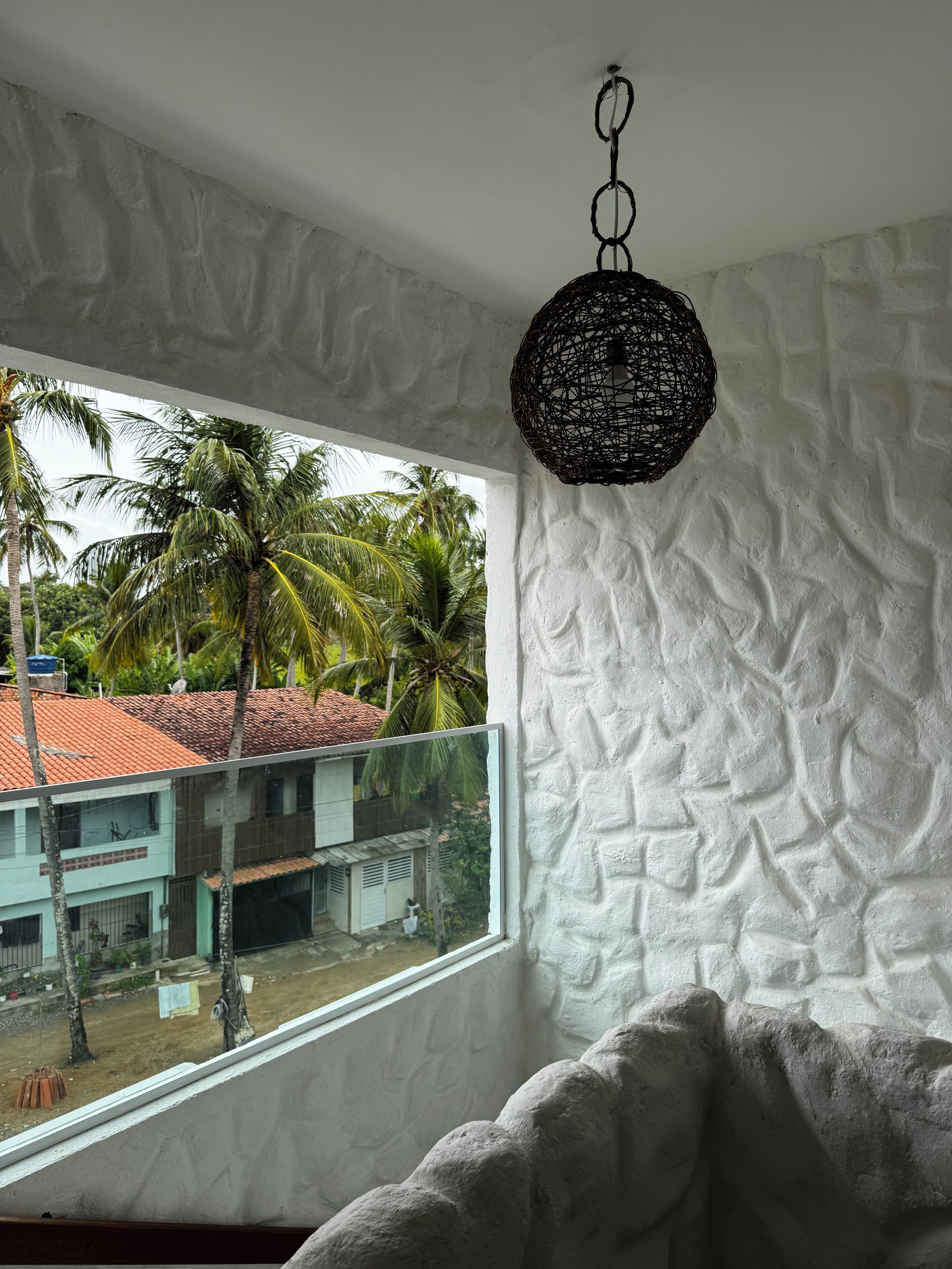 A room with a textured wall, a hanging light fixture, and a view of a building with a red roof and palm trees outside.