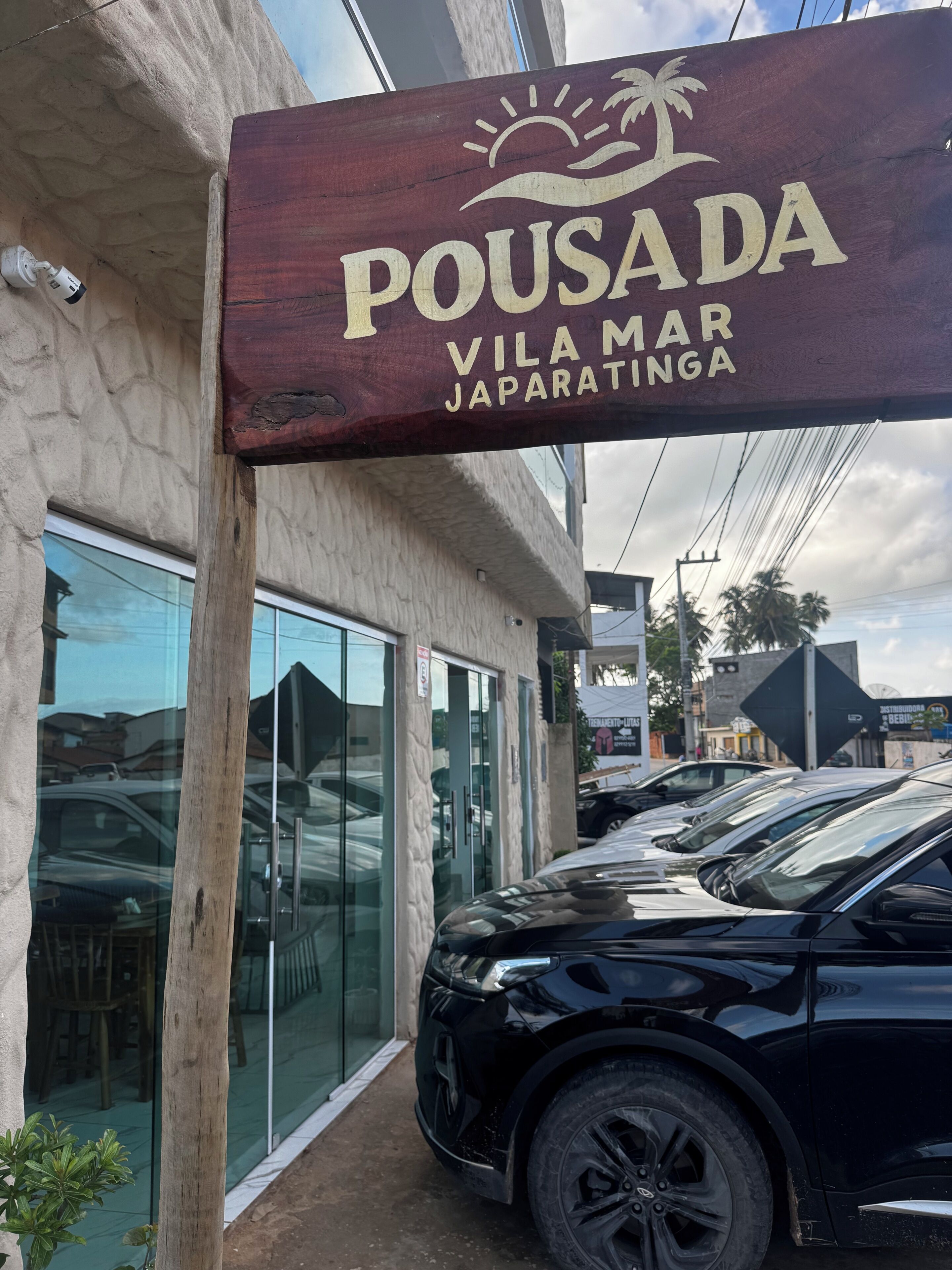A sign for 'Pousada Vila Mar Japaratinga' with a modern building facade and parked cars.