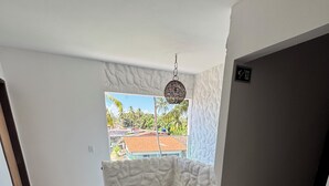 A room with a potted plant on a wooden table, a window with a view of buildings and palm trees, and a textured wall. - Pousada Vila Mar Japaratinga (Japaratinga)