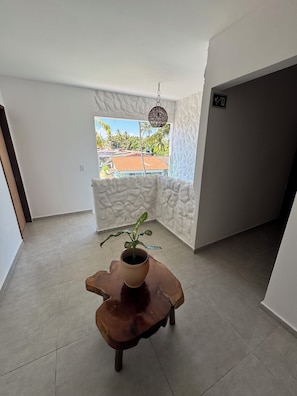 Habitació amb una planta en test, una taula de fusta, una finestra amb vista a edificis i palmeres, i una paret amb textura.