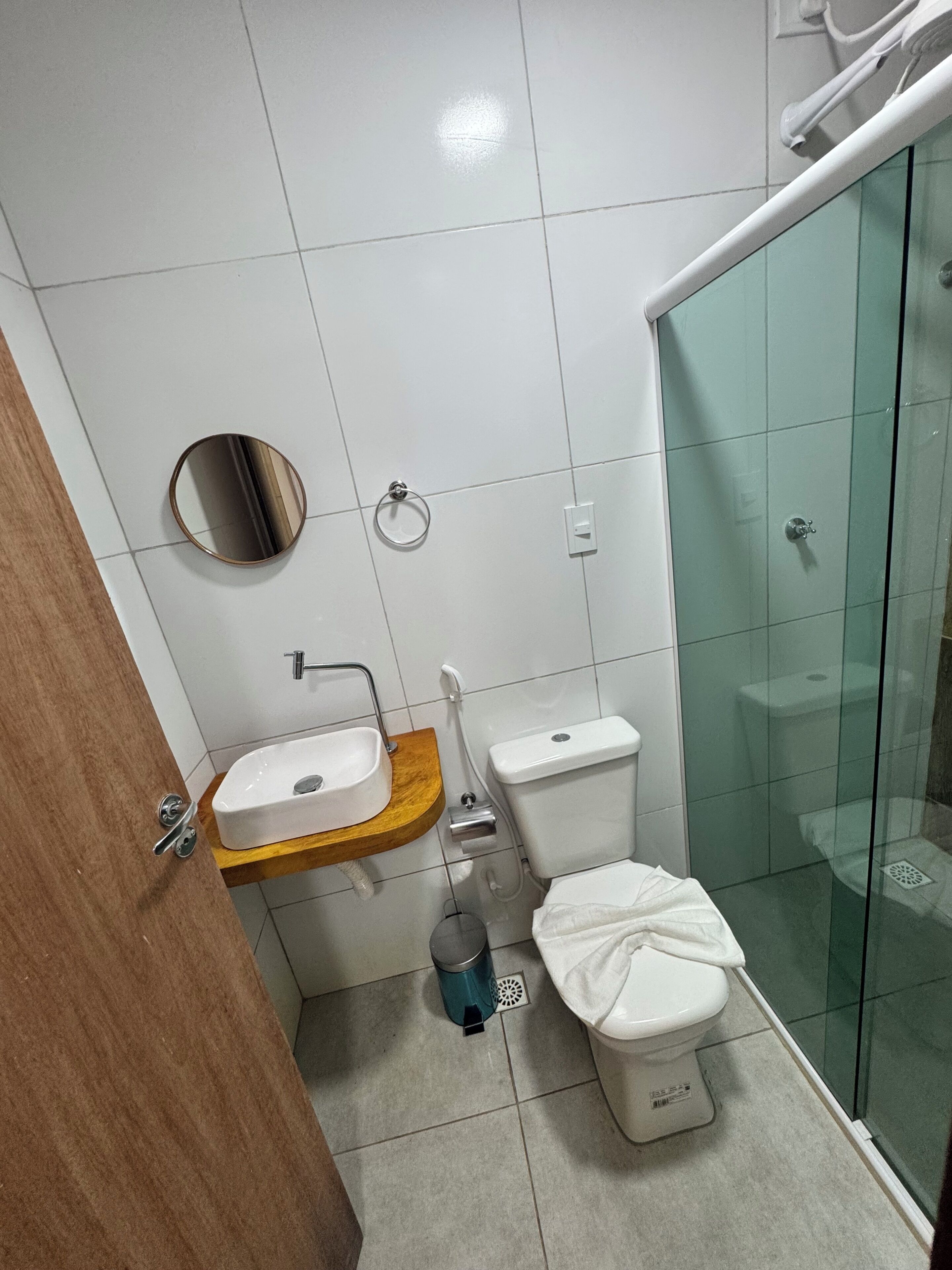 Een badkamer met een houten wastafelmeubel, een ronde spiegel, een toilet en een douchecabine met glazen wanden.