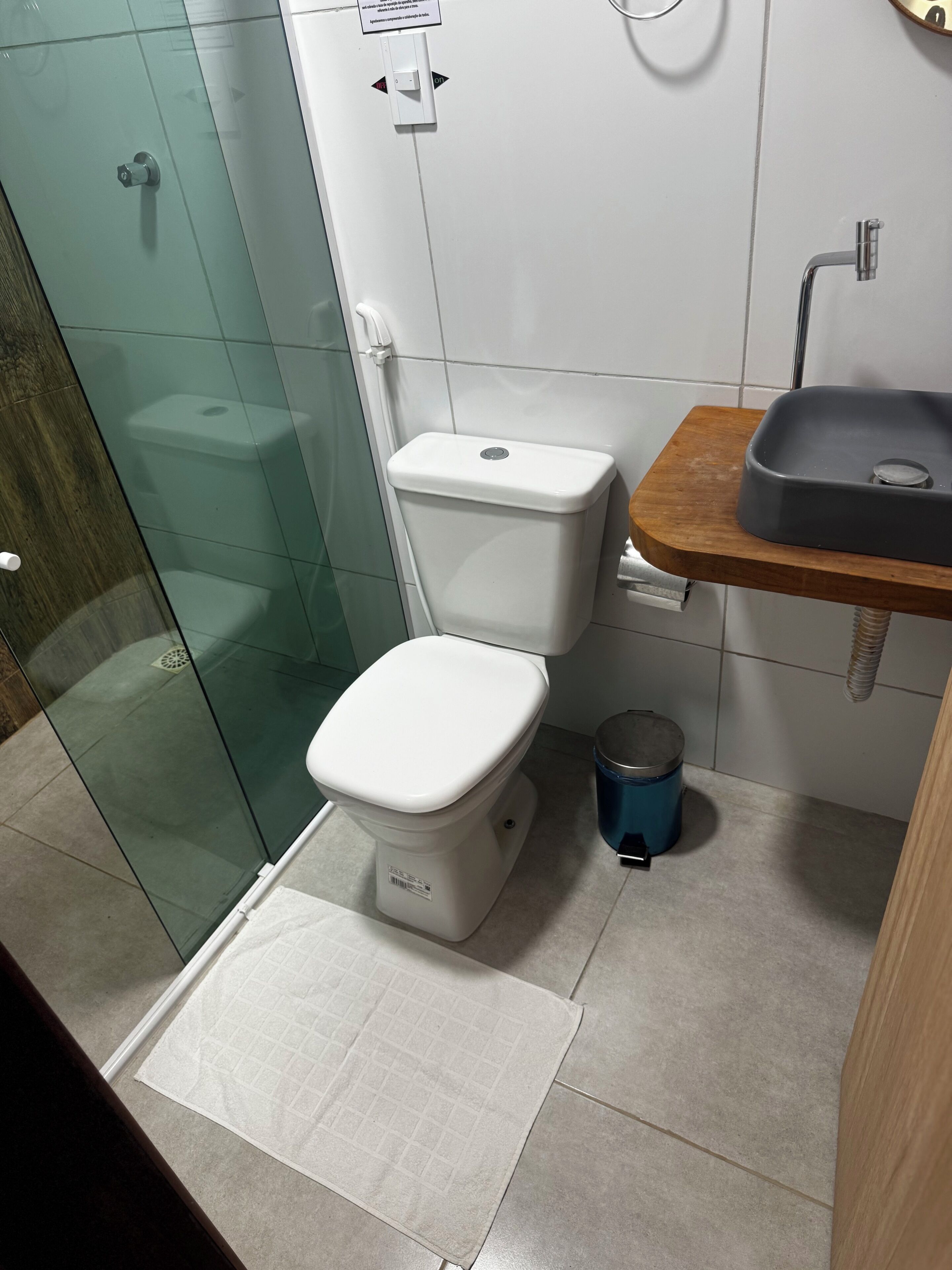 Un baño moderno con cabina de ducha de vidrio, inodoro blanco, tocador de madera con lavamanos y un cubo de basura azul.