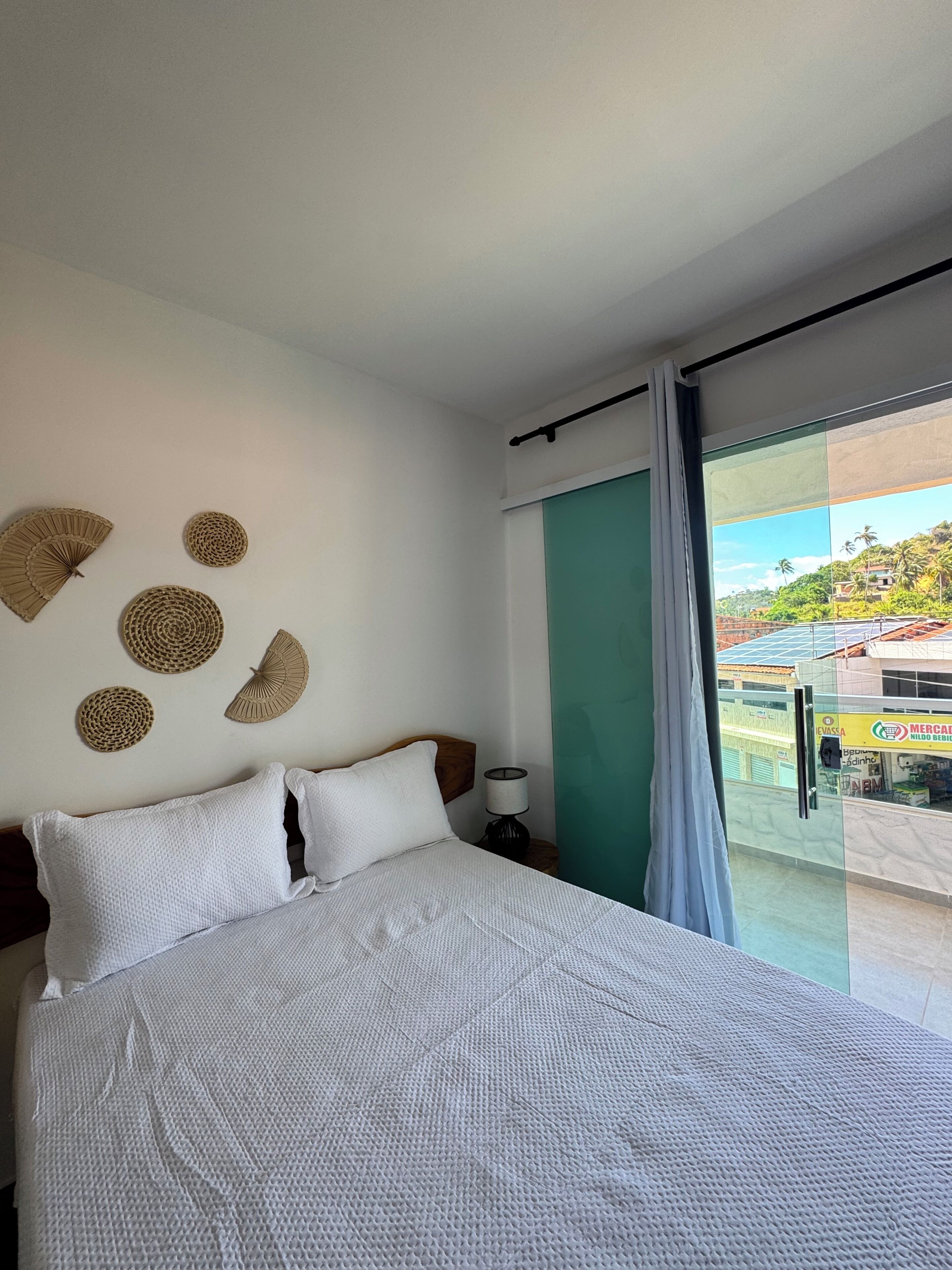 A hotel room with a bed, a nightstand, a wall-mounted lamp, and a view of the outside.