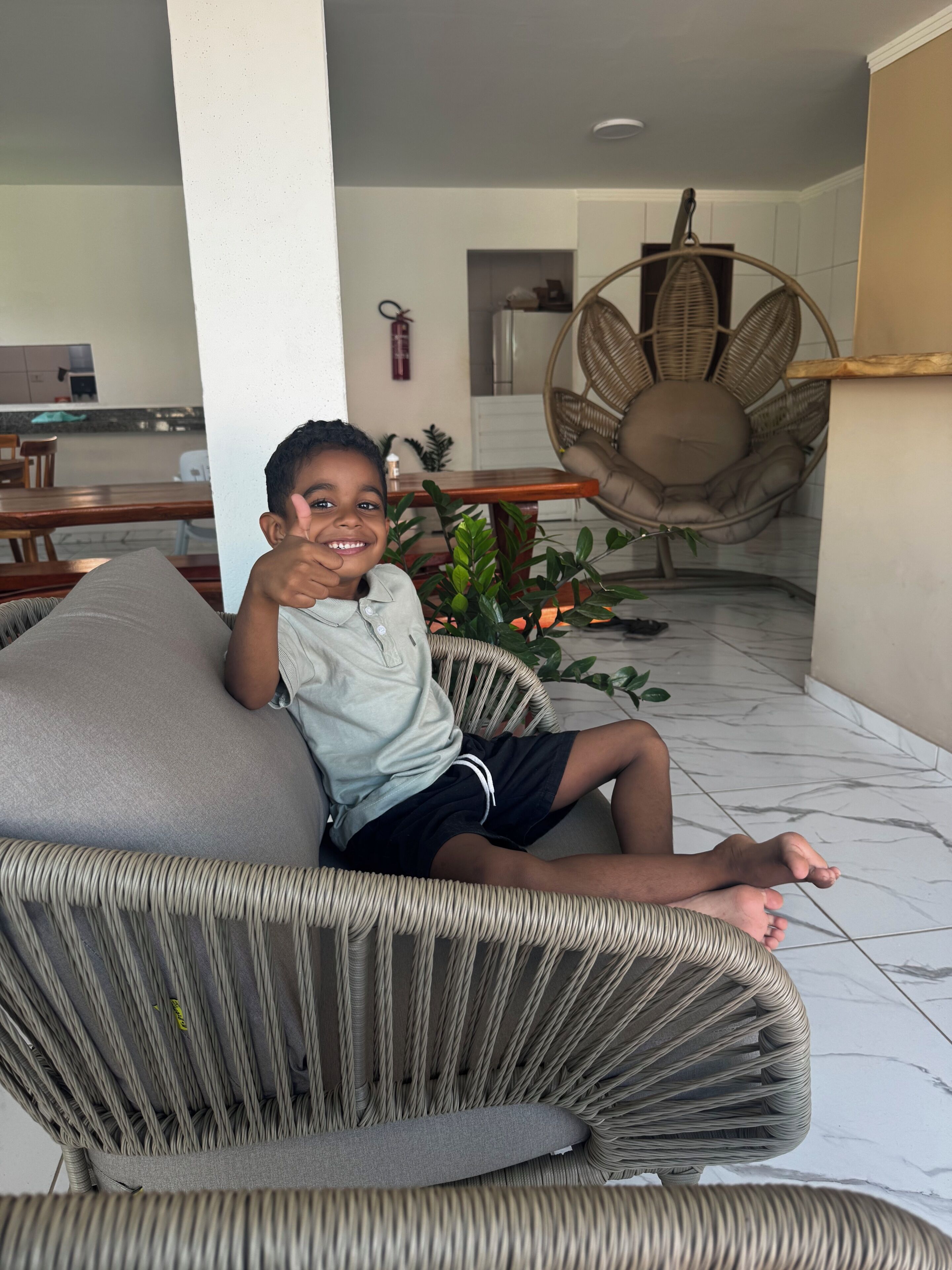 A child sitting on a wicker chair with a cushion, in a room with a tiled floor and a hanging chair in the background.