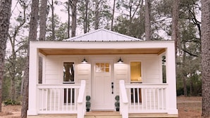 A small, white cabin with a wooden porch, two windows, and a door.