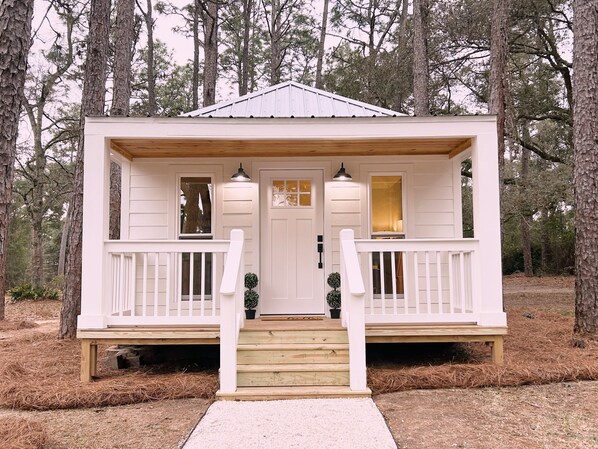 Una cabaña pequeña y blanca con un porche de madera, dos ventanas y una puerta.