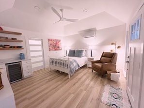 Un dormitorio con cama, una silla, una mesita de noche, una lámpara, un ventilador de techo y una ventana.
