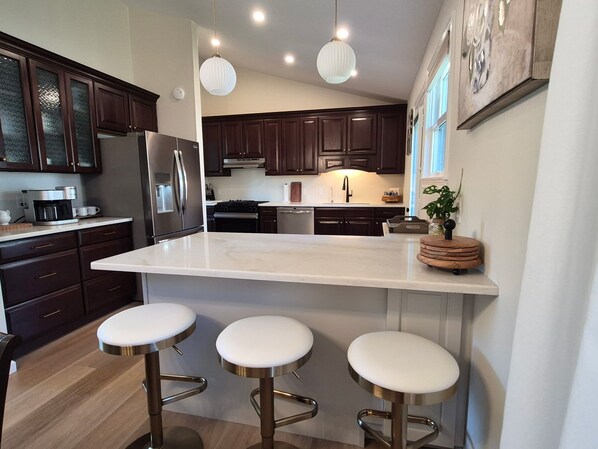 A modern kitchen with dark wood cabinets, a stainless steel refrigerator, a white marble island with bar stools, and pendant lighting.