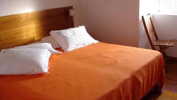 Een bed met een houten hoofdbord, witte kussens en een oranje sprei in een kamer met een houten stoel en een raam.