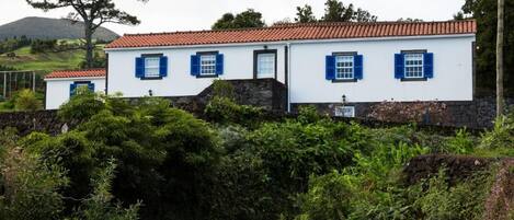 A two-story building with a red-tiled roof and white walls, surrounded by lush greenery and a stone wall.