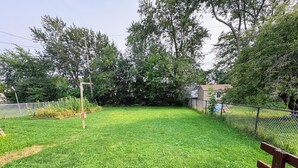 A grassy backyard with a wooden bench, a metal fence, and a house in the background.