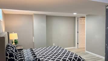 Quarto com cama, mesa de cabeceira, abajur e porta para outro cômodo.